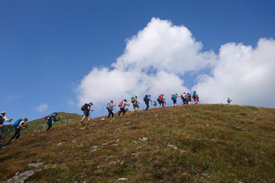 Eine Gruppe von Wanderern erklimmt einen Hügel unter einem blauen Himmel mit weißen Wolken. Die Landschaft ist grün und weitläufig. | © Weges OG
