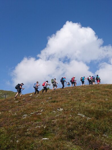 A group of hikers is climbing a hill under a blue sky with white clouds. The landscape is green and expansive. | © Weges OG