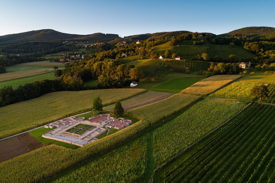 Eine weitläufige Landschaft mit sanften Hügeln und Weinbergen. In der Mitte sind die Ruinen einer ehemaligen Siedlung zu sehen. | © Oststeiermark Tourismus