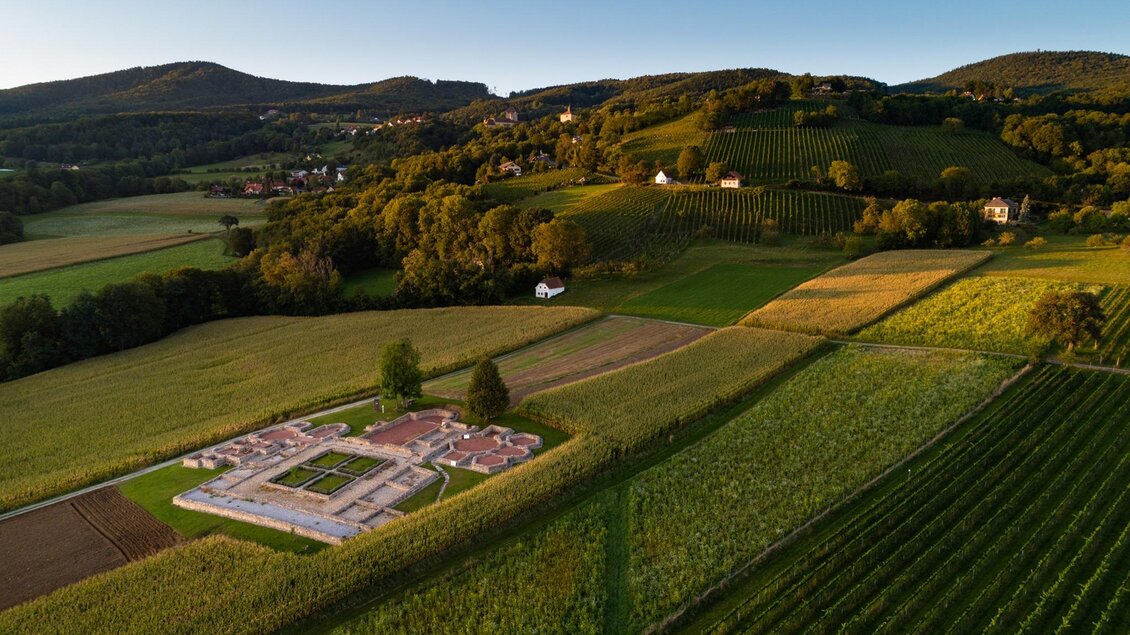 Eine weitläufige Landschaft mit sanften Hügeln und Weinbergen. In der Mitte sind die Ruinen einer ehemaligen Siedlung zu sehen. | © Oststeiermark Tourismus