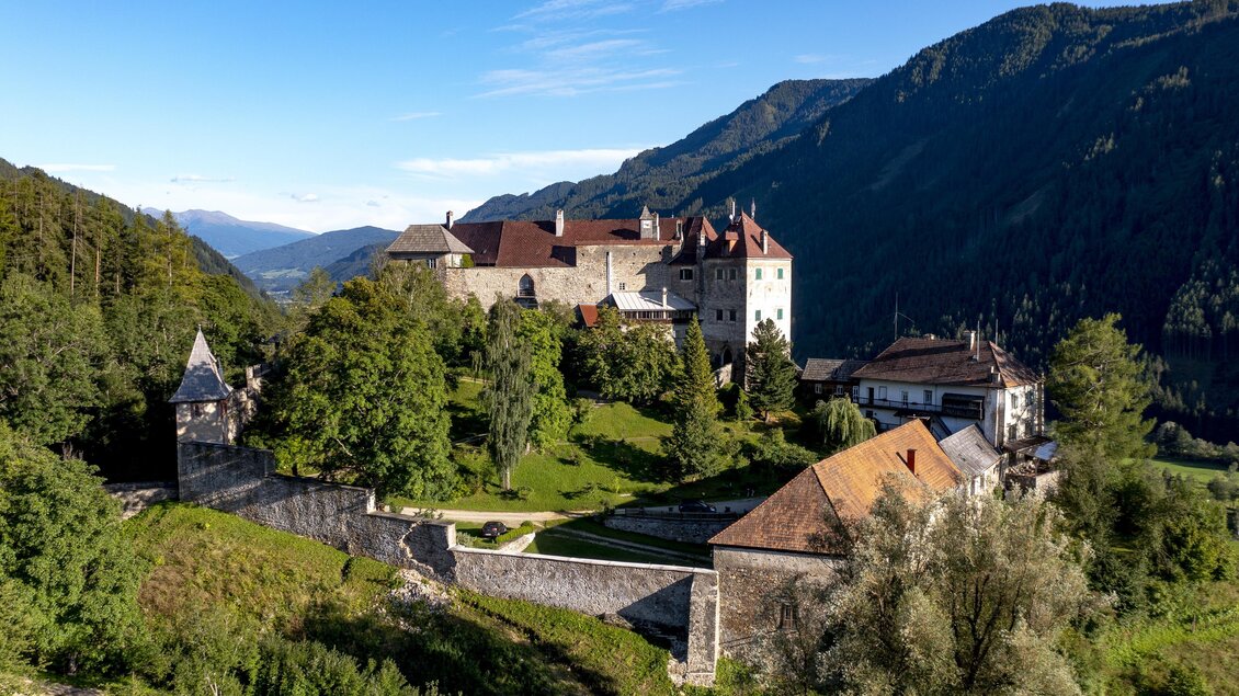 Eine beeindruckende Burg auf einem Hügel, umgeben von grünen Bäumen und Bergen. Der Himmel ist klar und sonnig. | © Tom Lamm