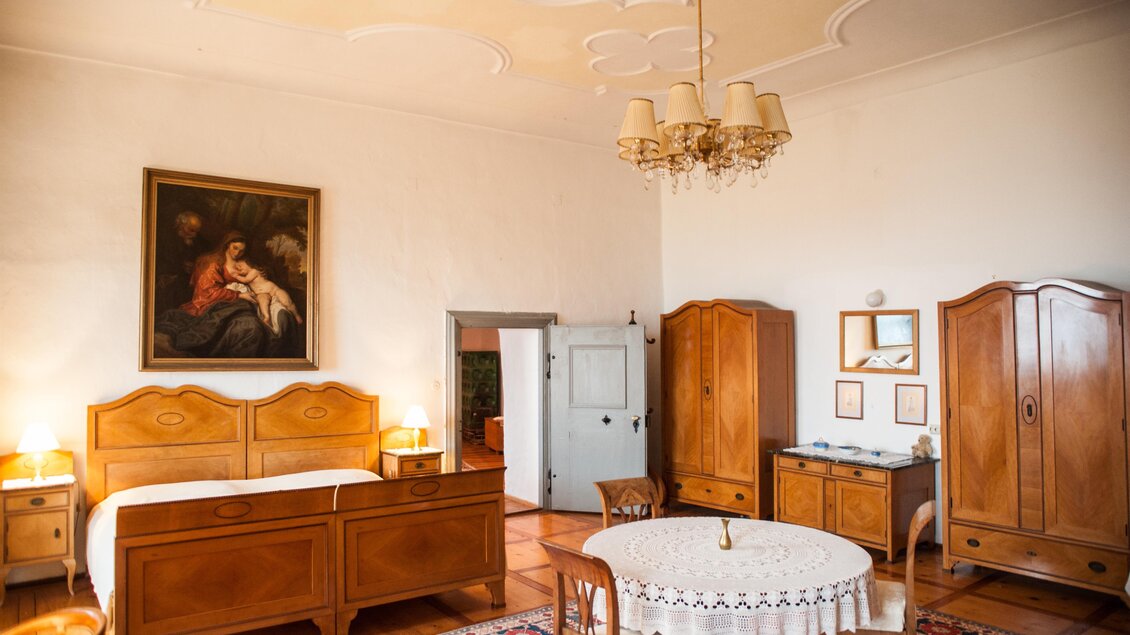 Ein großzügiges Zimmer mit Holzmöbeln und einem eleganten Kronleuchter. An der Wand hängt ein Gemälde, und es gibt einen kleinen Tisch mit Stühlen. | © Fam. Steiner