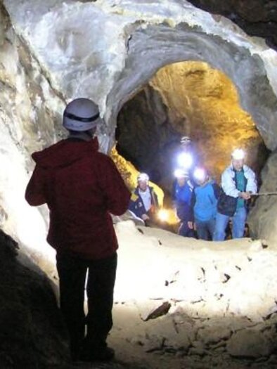 Eine Gruppe von Menschen mit Helmen wandert durch eine Höhle. Das Licht ihrer Lampen erhellt den engen Tunnel.