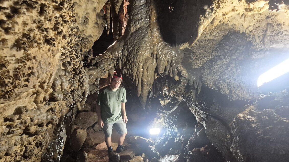 Eine Person steht in einer Höhle mit Stalaktiten und Stalagmiten. Die Szenerie ist schwach beleuchtet, mit einigen Lichtquellen, die den Weg erhellen. | © Andrea Stelzer