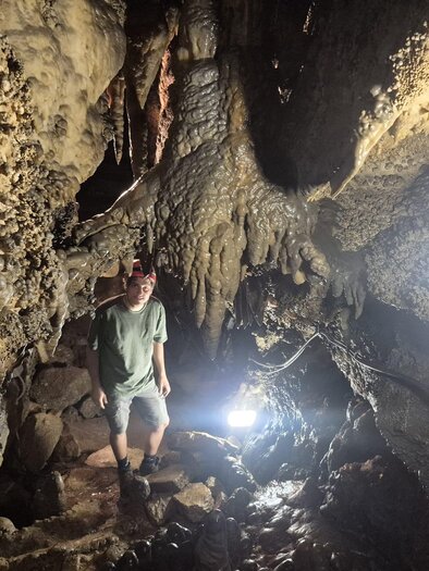 A person is standing in a cave with stalactites and stalagmites. The scene is dimly lit, with some light sources illuminating the path. | © Andrea Stelzer