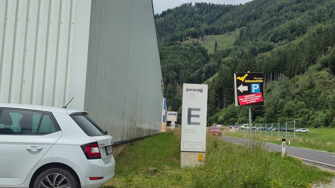Ein Parkplatz mit einem weißen Auto und einem Schild. Im Hintergrund sind Berge und ein typisches ländliches Gelände zu sehen. | © Andrea Stelzer