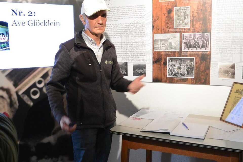 Ein Mann steht vor einer Wand mit historischen Fotos und Texten in einem Museum. Er erklärt Informationen zu einem Thema, während ein Tisch mit Dokumenten im Vordergrund sichtbar ist. | © Maria Krause