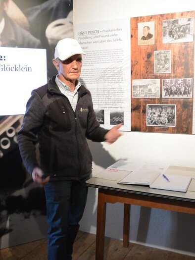 Ein Mann steht vor einer Wand mit historischen Fotos und Texten in einem Museum. Er erklärt Informationen zu einem Thema, während ein Tisch mit Dokumenten im Vordergrund sichtbar ist. | © Maria Krause