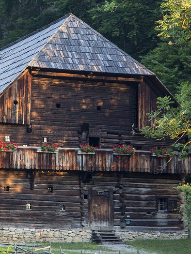 A rustic wooden house surrounded by trees. The balcony is decorated with flowers and the roof has a distinctive shape. | © Freilichtmuseum Stübing