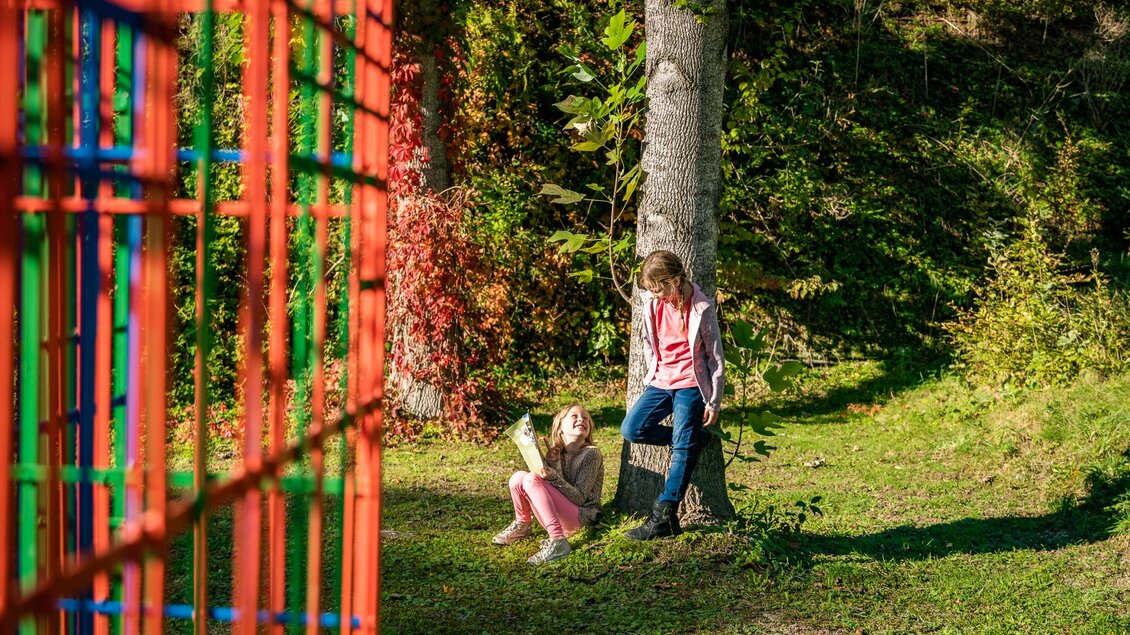 Zwei Kinder sitzen und stehen bei einem Baum im Skulpturenpark neben einer bunten Kunstinstallation. | © Sabina Hörter, o.T.; Region Graz - Mias Photoart