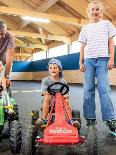 Eine fröhliche Gruppe von Kindern fährt mit Spielzeugfahrzeugen in einer hellen Indoor-Spielumgebung. Ein Erwachsener steht hinter den fahrbaren Geräten und lächelt. | © www.zloam.at
