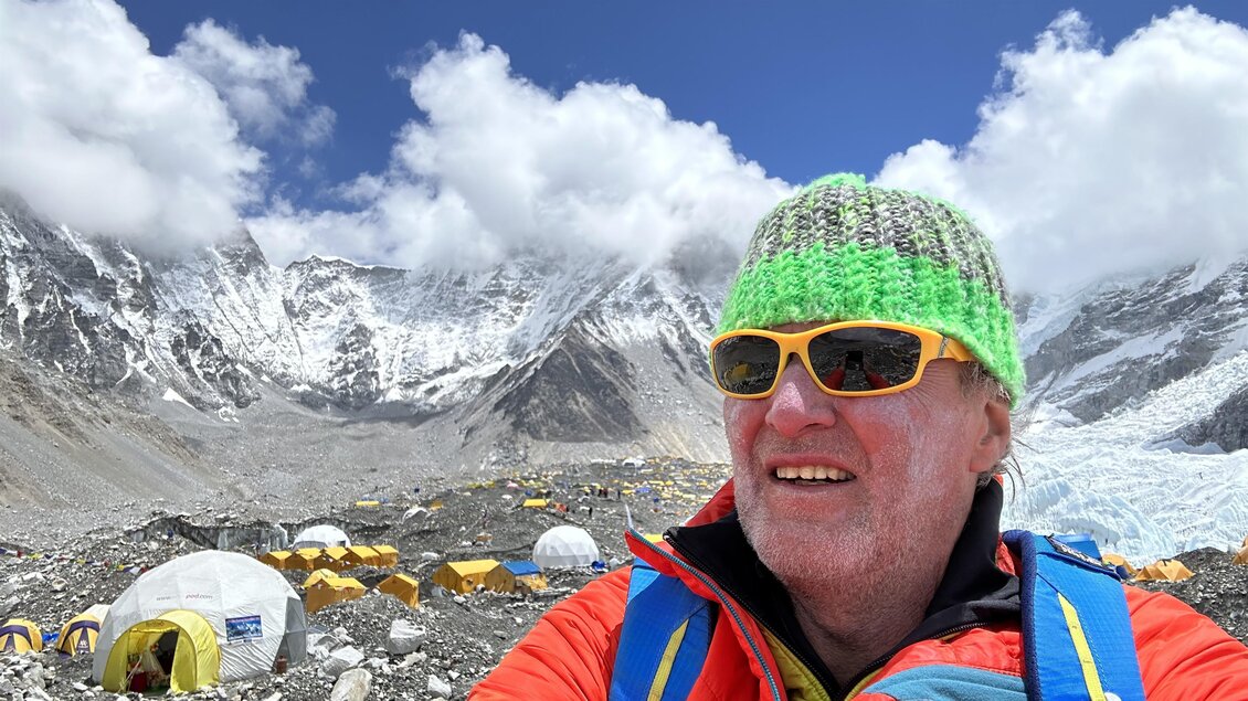 Ein Bergsteiger mit buntem Hut steht vor dem Mount Everest und lächelt. Im Hintergrund sind schneebedeckte Berge und Lagerzelte sichtbar. | © Helmut Pichler