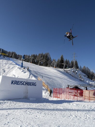 A skier jumps over a jump in the snow. In the background, trees and a blue sky can be seen. | © TVB Murau