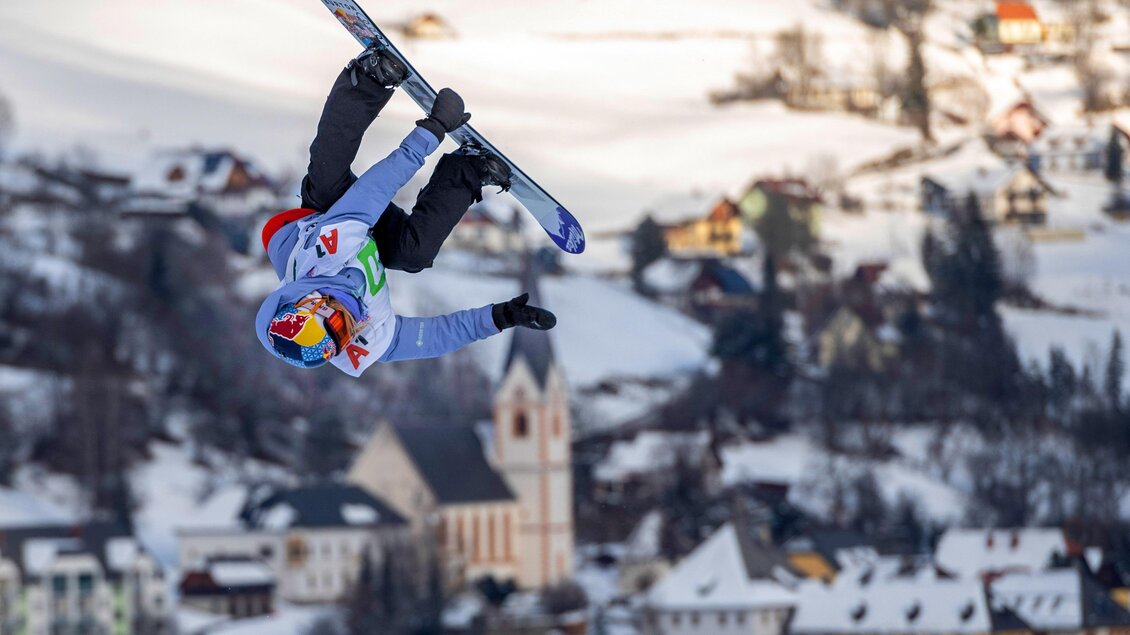 Ein Snowboardfahrer führt einen Trick in der Luft aus. Hintergrund mit schneebedeckten Hügeln und kleinen Häusern. | © TVB Murau