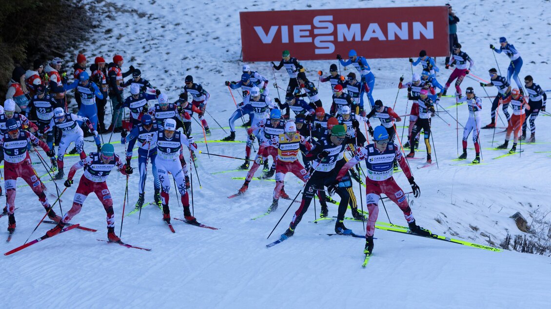 Eine Gruppe von Skilangläufern startet ein Rennen auf verschneitem Gelände. Im Hintergrund ist ein Viesmann-Werbeschild zu sehen.