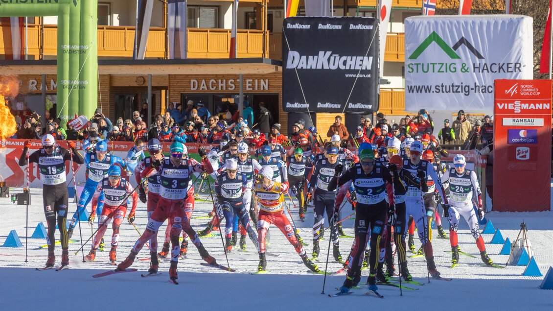 Ein Wettkampf im Langlauf mit vielen Athleten auf Schnee. Im Hintergrund sind Gebäude und Sponsorenbanner zu sehen.