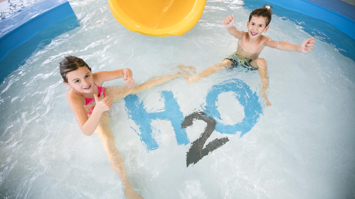 Zwei Kinder spielen im Wasser, lächelnd und voller Freude. Im Hintergrund ist eine gelbe Rutsche und ein großes "H2O" im Wasser zu sehen. | © TV BW