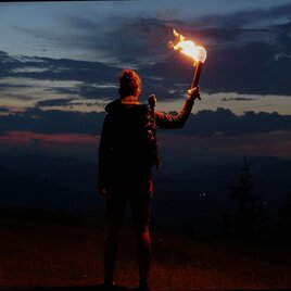 Person with a burning torch on a mountain meadow near Weinitzen in front of dark mountain silhouettes at dusk. | © SurWhyve