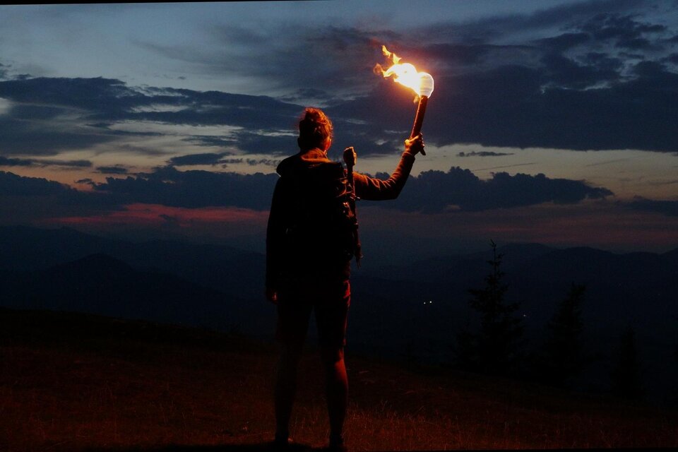 Person mit brennender Fackel auf einer Bergwiese bei Weinitzen vor dunklen Bergsilhouetten in der Abenddämmerung. | © SurWhyve