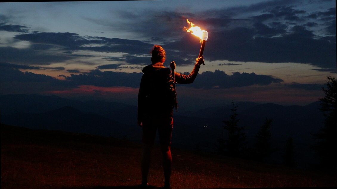 Person mit brennender Fackel auf einer Bergwiese bei Weinitzen vor dunklen Bergsilhouetten in der Abenddämmerung. | © SurWhyve