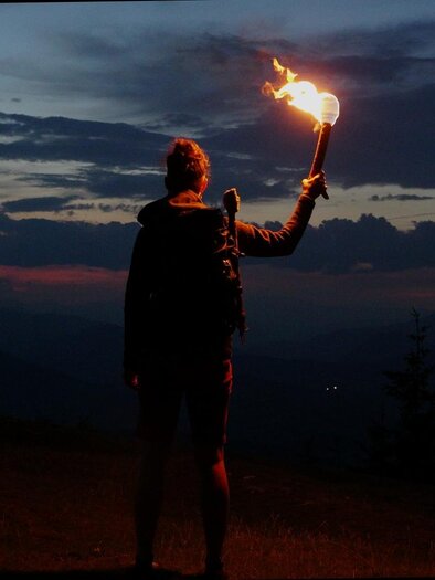 Person mit brennender Fackel auf einer Bergwiese bei Weinitzen vor dunklen Bergsilhouetten in der Abenddämmerung. | © SurWhyve