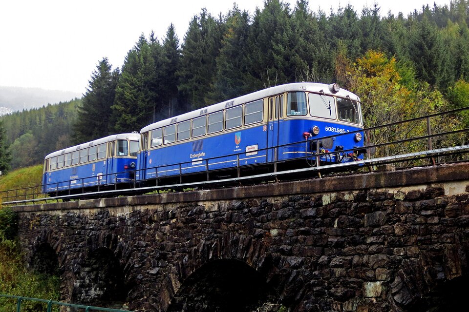 Ein blauer Zug fährt über eine alte Steinbrücke in einer bewaldeten Landschaft. Der Himmel ist bewölkt und die Natur um ihn herum ist grün. | © Verein Erzbergbahn