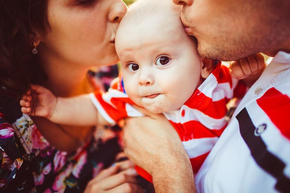 Ein Baby wird liebevoll von seinen Eltern geküsst. Es trägt ein gestreiftes Shirt und hat einen neugierigen Gesichtsausdruck. | © Bild: Freepik