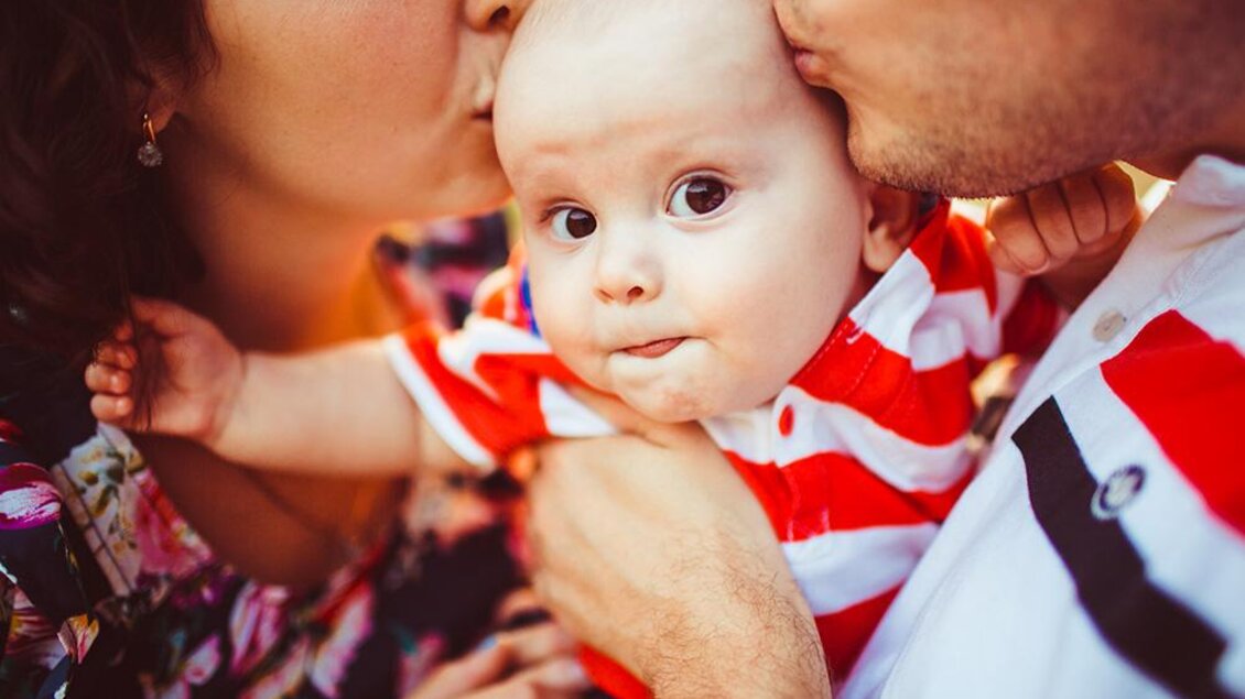 Ein Baby wird von seinen Eltern geküsst. Es trägt ein gestreiftes Oberteil und hat einen süßen Ausdruck. | © Bild: Freepik