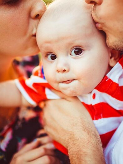 Ein kleines Baby in einem gestreiften Hemd wird von seinen Eltern umarmt und geküsst. Die Szene strahlt Wärme und Liebe aus. | © Bild: Freepik