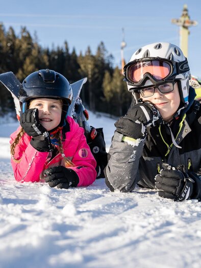Két gyerek fekszik a hóban és mosolyog. Síruhát és sisakot viselnek, míg a háttérben fák és egy sífelvonó látható. | © Tourismusverband Oststeiermark