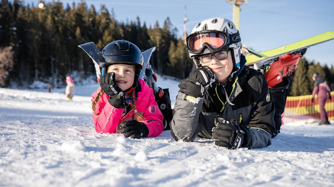 Zwei Kinder liegen im Schnee, eines in pinker Skijacke und das andere in schwarzer Jacke. Sie tragen Skihelme und schauen lächelnd zur Kamera. | © Tourismusverband Oststeiermark