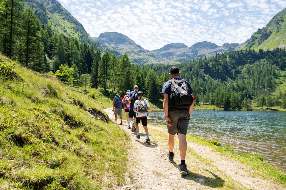 Eine Gruppe von Wanderern geht entlang eines Weges an einem klaren See, umgeben von Bergen und Bäumen. Der Himmel ist mit Wolken bedeckt und die Landschaft ist üppig grün. | © TVB/Laura Passenbrunner