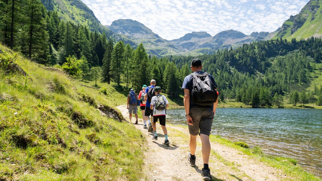 Eine Gruppe von Wanderern geht entlang eines Weges an einem klaren See, umgeben von Bergen und Bäumen. Der Himmel ist mit Wolken bedeckt und die Landschaft ist üppig grün. | © TVB/Laura Passenbrunner
