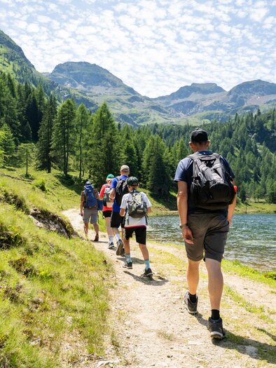 Eine Gruppe von Wanderern geht entlang eines Weges an einem klaren See, umgeben von Bergen und Bäumen. Der Himmel ist mit Wolken bedeckt und die Landschaft ist üppig grün. | © TVB/Laura Passenbrunner