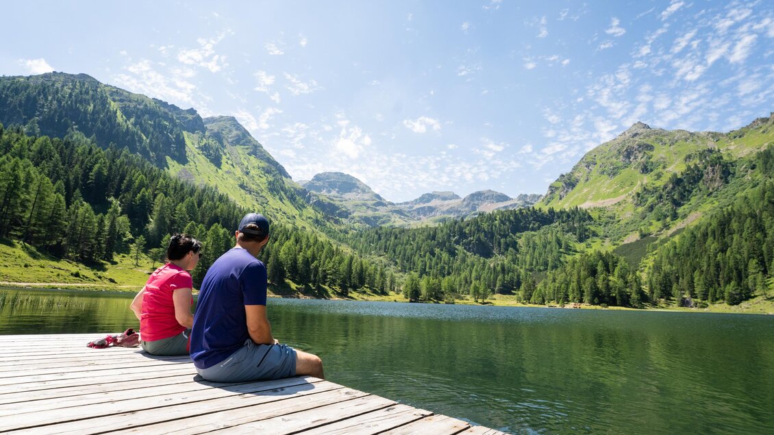 Ein Paar sitzt auf einem Holzsteg am See und schaut auf die umliegenden Berge. Die Landschaft ist grün und sonnig, mit einem klaren Himmel. | © TVB/Laura Passenbrunner