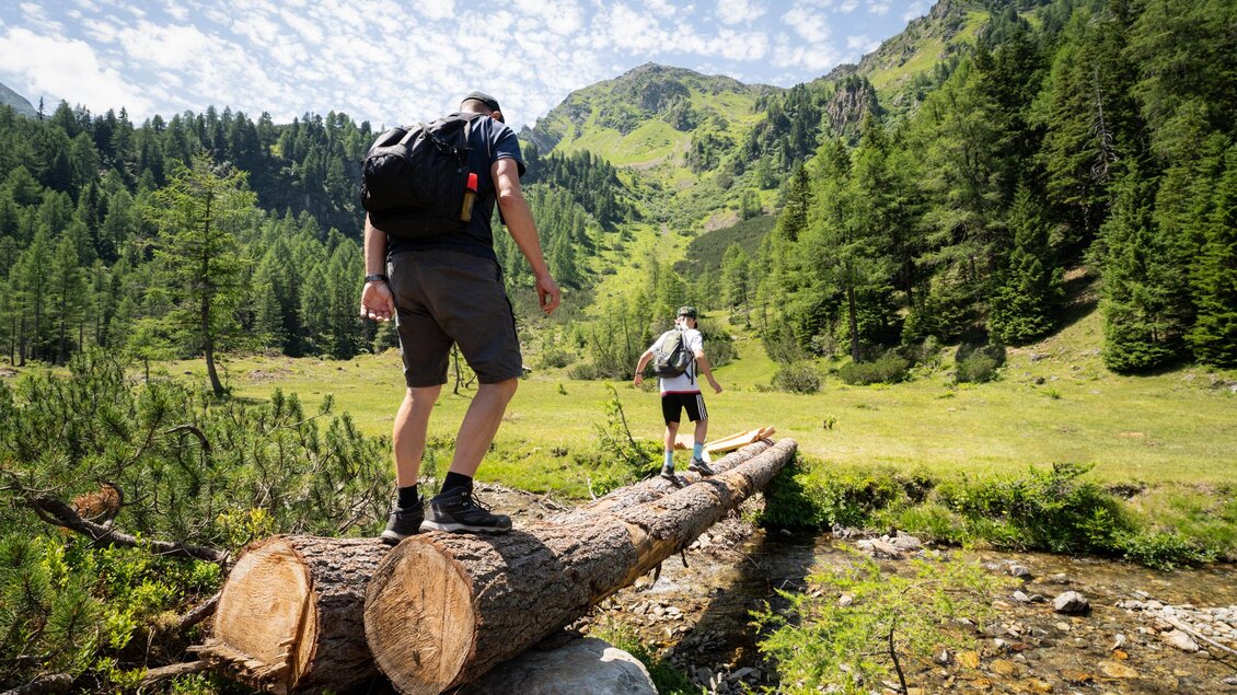 Zwei Wanderer überqueren einen Baumstamm über einen klaren Bach in einer malerischen Berglandschaft. Die Umgebung ist grün, umgeben von hohen Bäumen und Bergen. | © TVB/Laura Passenbrunner