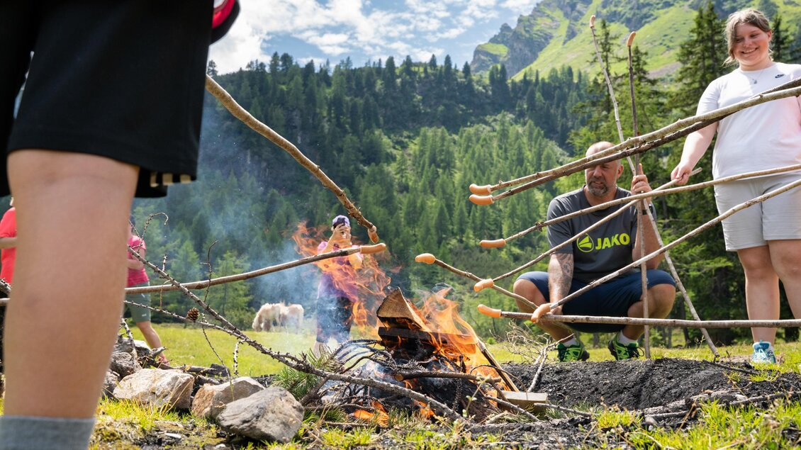 Eine Gruppe von Menschen grillt am Lagerfeuer in einer idyllischen Berglandschaft. Im Hintergrund sind grüne Bäume und eine klare Himmel zu sehen. | © TVB/Laura Passenbrunner
