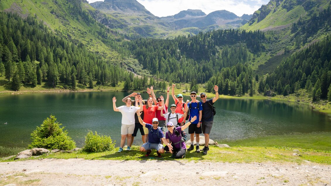 Eine Gruppe von Menschen steht am Ufer eines Sees, umgeben von grünen Bergen und Bäumen. Sie lachen und freuen sich, während sie die Natur genießen. | © TVB/Laura Passenbrunner