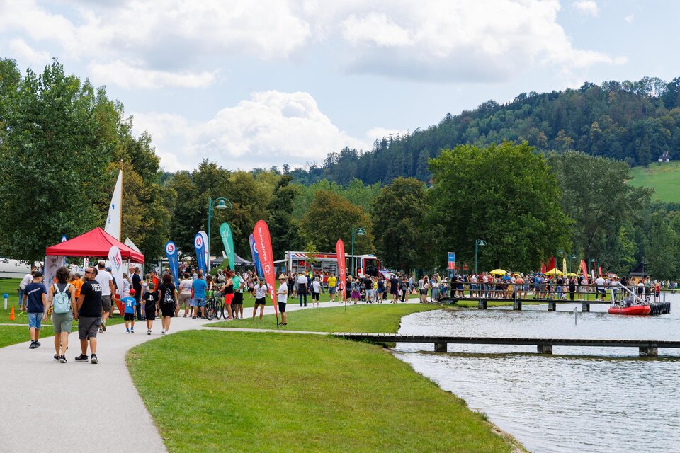 Eine belebte Promenade am See mit vielen Besuchern und bunten Fahnen. Im Hintergrund sind Bäume und ein Hügel sichtbar. | © Woche Familienfest