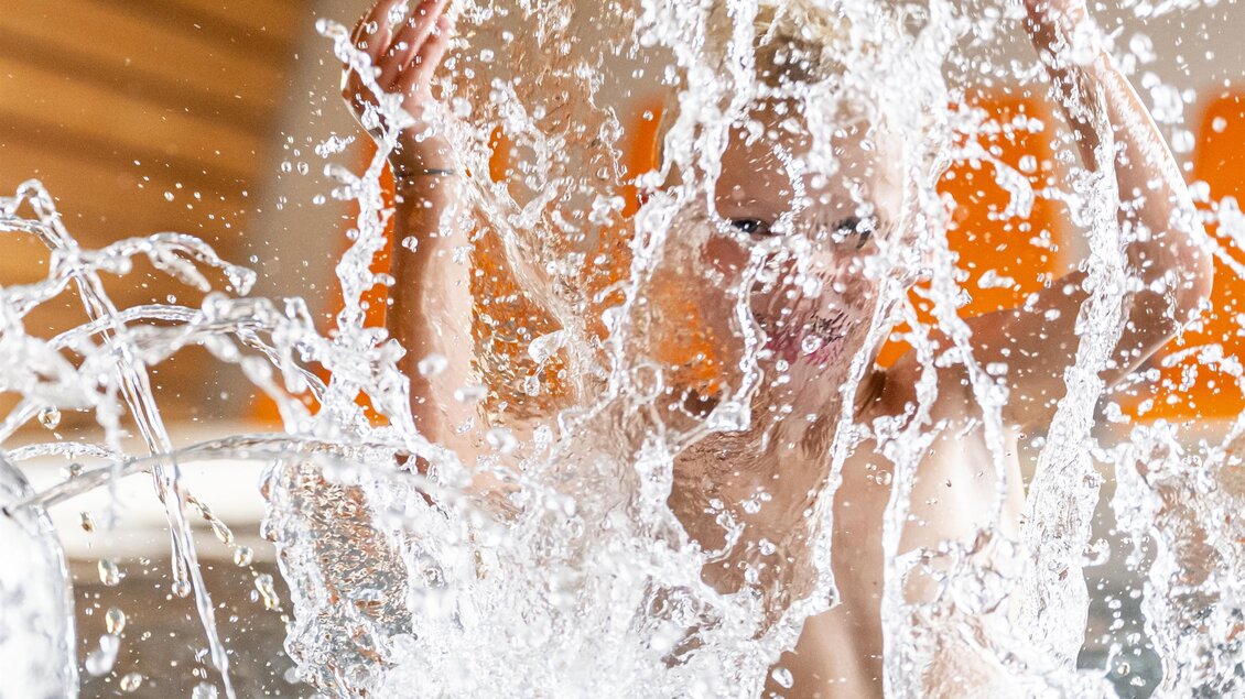 Ein Kind spritzt fröhlich im Wasser und erzeugt Spritzwasser. Im Hintergrund sind orangefarbene Liegen zu sehen. | © Therme Aqualux