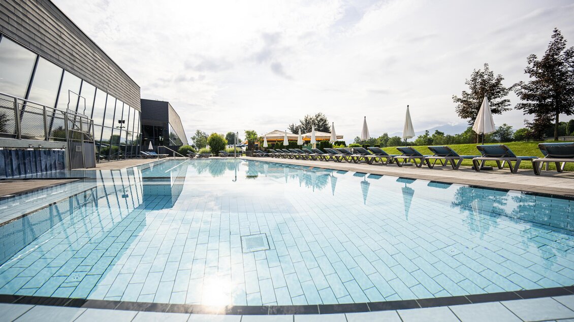 Ein schöner Pool mit klarem Wasser und modernen Liegen. Im Hintergrund zeigt sich eine grüne Landschaft mit Sonnenschirmen. | © Therme Aqualux