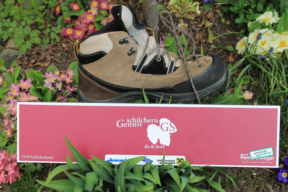Ein Wanderschuh steht auf einem bunte Blumenbeet. Darunter befindet sich ein rotes Schild mit einer Beschreibung. | © TV Südsteiermark - Michi Lang