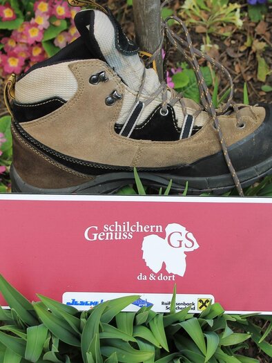 A hiking shoe is standing on a colorful flower bed. Below it is a red sign with a description. | © TV Südsteiermark - Michi Lang