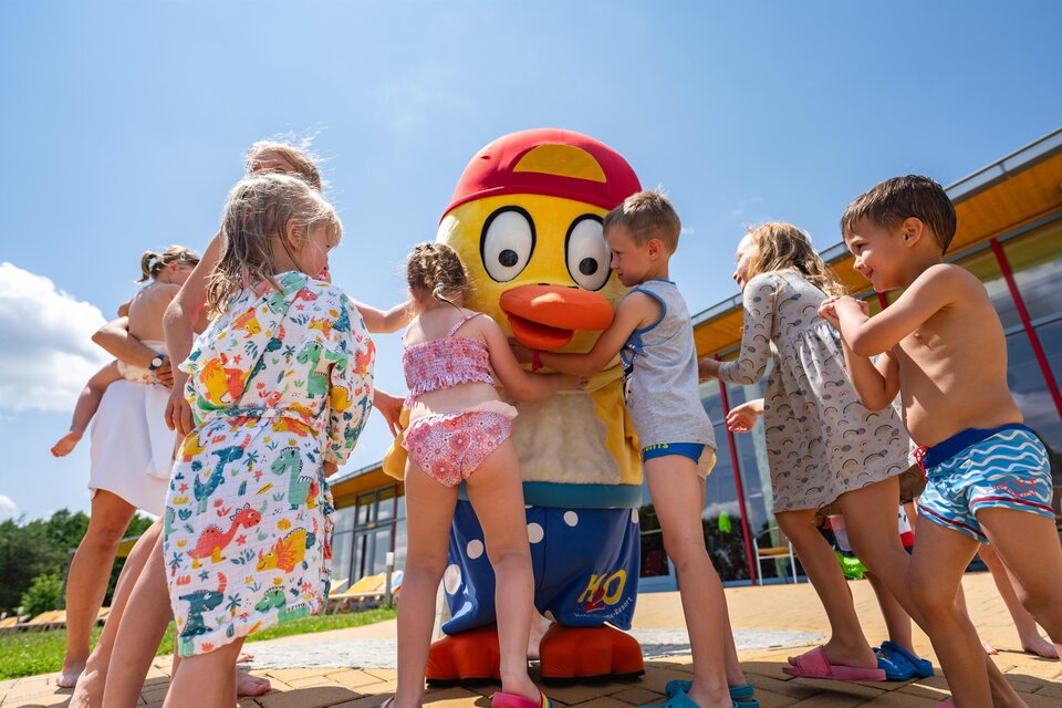 Eine Gruppe von Kindern umarmt eine große, lustige Entenfigur. Es ist ein sonniger Tag und die Kinder tragen Bademode. | © H2O-Hoteltherme GmbHmoving.stills
