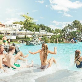A sunny pool area with happy children playing in the water. In the background, water slides and green trees can be seen. | © Thermenresort Loipersdorf