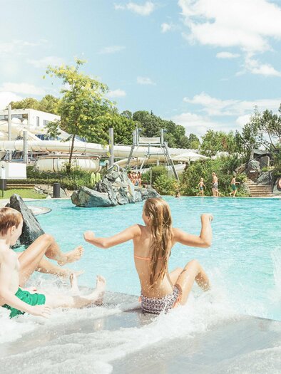 Ein sonniger Poolbereich mit fröhlichen Kindern, die im Wasser spielen. Im Hintergrund sind Wasserrutschen und grüne Bäume zu sehen. | © Thermenresort Loipersdorf