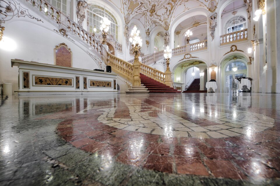 Ein beeindruckender Innenraum mit einer prunkvollen Treppe und kunstvollen Deckenverzierungen. Der polierte Boden reflektiert die elegante Architektur. | © Opernhaus Graz