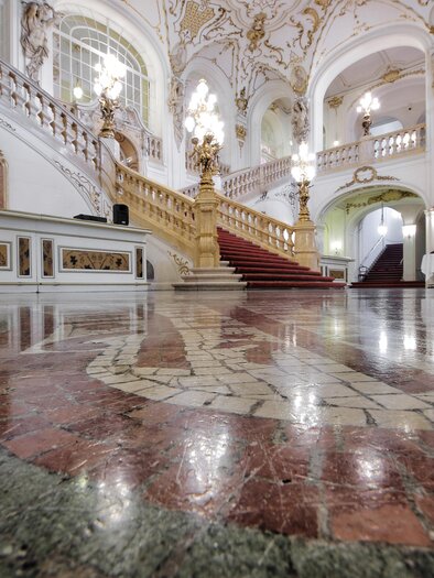 An impressive interior with an ornate staircase and elaborate ceiling decorations. The polished floor reflects the elegant architecture. | © Opernhaus Graz