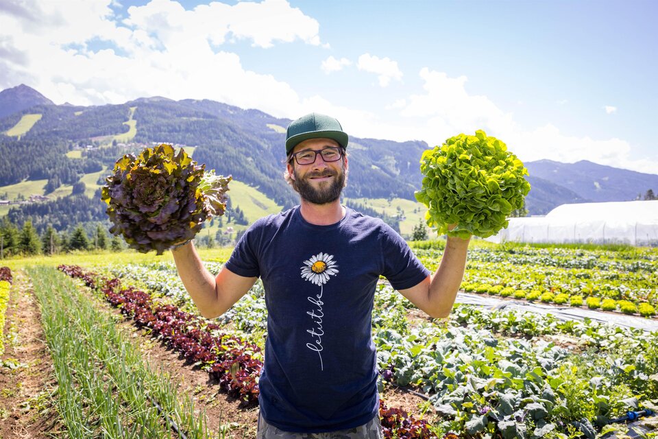 Ein lächelnder Mann steht auf einem Feld und hält in jeder Hand einen Kopfsalat. Im Hintergrund sind Berge und grüne Pflanzen zu sehen. | © Michael Windberger