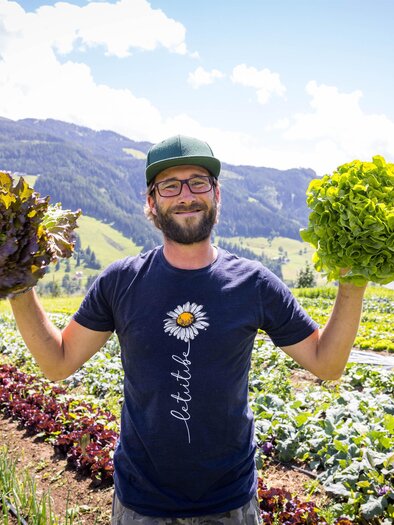 Ein lächelnder Mann steht auf einem Feld und hält in jeder Hand einen Kopfsalat. Im Hintergrund sind Berge und grüne Pflanzen zu sehen. | © Michael Windberger